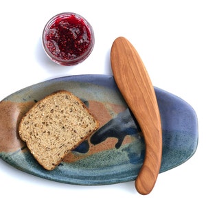 May include: A blue and brown ceramic serving dish with a wooden butter knife and a slice of bread. A small glass jar of red raspberry jam is on the side.