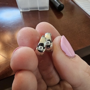 May include: A pair of silver hoop earrings with a small, clear gemstone set into each earring. The earrings are held in a person's hand, with a light pink nail polish visible.