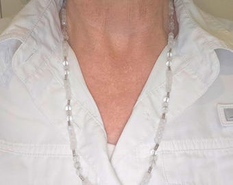 Pode incluir: Um colar longo e delicado com contas claras e facetadas e pequenos detalhes metálicos prateados. O colar é usado sobre uma camisa branca com gola, criando um visual simples, mas elegante. As contas parecem ser semitransparentes.