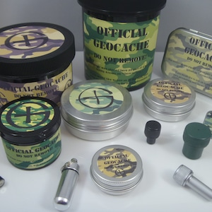 May include: A collection of various geocaching containers including black and silver plastic jars, metal tins, and small cylindrical tubes. Each item features camouflage patterns and labels that read OFFICIAL GEOCACHE DO NOT REMOVE.