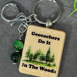 May include: A rectangular wooden keychain featuring a printed forest scene and the black text Geocachers Do It In The Woods. The keychain includes a silver metal split ring, a lobster clasp, and a dangling charm with two green tiger eye beads.