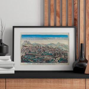 May include: Framed vintage print of a city landscape with mountains and a blue sky. The artwork is in a black frame, displayed on a black shelf with decorative vases and a stack of books. The color palette includes blues, greens, and browns.