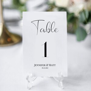 Op de afbeelding: Witte tafelnummerkaart met zwarte kalligrafie die "Tafel 1" en de tekst "Jennifer & Matt 05.22.2022" in zwart zegt.