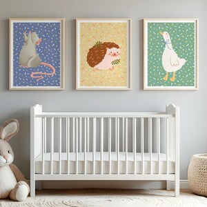 Pode incluir: Três impressões de arte emolduradas com ilustrações de animais: um rato cinzento, um ouriço-cacheiro castanho e um ganso branco. As impressões estão acima de um berço branco num quarto de criança. A arte tem um estilo infantil e fantasioso.