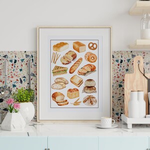 May include: A framed watercolor print featuring various types of bread, including loaves, rolls, and sandwiches. The artwork showcases a variety of baked goods in shades of brown and beige, with a pretzel and wheat stalks. The print is displayed on a kitchen counter.
