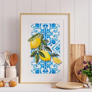Pode incluir: Uma impressão em aquarela emoldurada com limões em um galho com folhas verdes. O fundo apresenta um padrão de azulejos azuis e brancos. A obra de arte é exibida em uma moldura dourada, evocando um tema mediterrâneo.