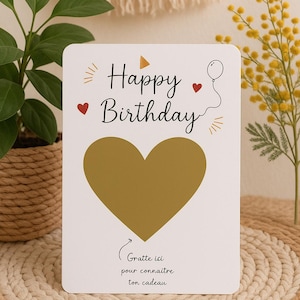 Peut inclure: Une carte d'anniversaire blanche avec l'inscription "Happy Birthday" en noir, une zone à gratter en forme de cœur doré et une illustration de ballon. La carte porte également l'inscription "Grattez ici pour connaître ton cadeau".
