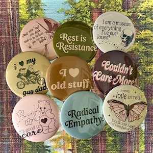 May include: A collection of round pinback buttons with various designs and text. The buttons feature phrases like "Rest is Resistance," "Couldn't Care More!", and "Radical Empathy!" Other buttons have illustrations of a skull, a butterfly, and a bear.