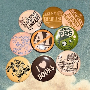 May include: A collection of nine round buttons with various designs and text. The buttons feature phrases like "Support Your Local Library," "Take Me Thrifting," and "I Love Books." Colors include pink, brown, yellow, and green.