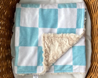 May include: A soft, blue and white checkered baby blanket with a plush, beige sherpa lining. The blanket is folded in half and is sitting in a wicker basket.