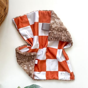 May include: A plush baby blanket with a checkerboard pattern in white and orange. The blanket has a soft, brown, faux fur trim. The label reads "Daily Myles".
