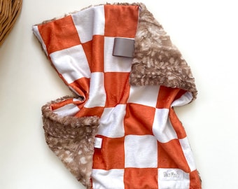 May include: A plush baby blanket with a checkerboard pattern in white and orange. The blanket has a soft, brown, faux fur trim. The label reads "Daily Myles".