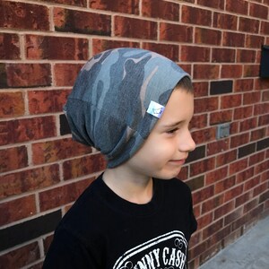 May include: A child wearing a gray and brown camouflage beanie with a white label. The beanie is made of a soft, stretchy fabric and has a slouchy fit.