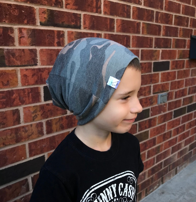 May include: A child wearing a gray and brown camouflage beanie with a white label. The beanie is made of a soft, stretchy fabric and has a slouchy fit.