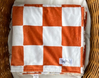 May include: A white and burnt orange checkered baby blanket with a "Daily Myles" label sewn on the bottom right corner.