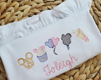 May include: A white shirt with ruffled sleeves featuring embroidered designs of a gold pretzel, a cupcake, three balloons, a black ice cream bar, and a waffle cone. The name Joleigh is embroidered in pink cursive script below the icons.