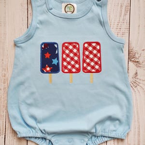 May include: Pale blue baby romper featuring three popsicle motifs. The left popsicle has a navy background with red and white stars. The other two have a red and white gingham pattern. The romper has snap closures.
