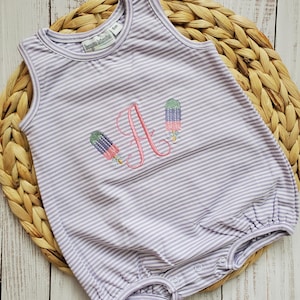 May include: A lavender and white striped baby romper with an embroidered floral design and the letter "A". The romper has a round neckline and snap closures at the bottom. The label reads "bugsy & kooks" and "6m".