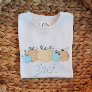 Puede incluir: Una camiseta blanca con un diseño bordado de cuatro calabazas en tonos azul claro, beige y amarillo, con hojas y enredaderas verdes. El nombre Jack está bordado en una fuente cursiva de color azul verdoso debajo de las calabazas.