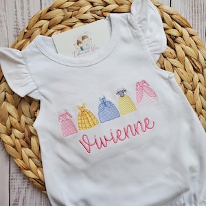 May include: White baby onesie with embroidered images of princess dresses and the name "Vivienne" in pink.