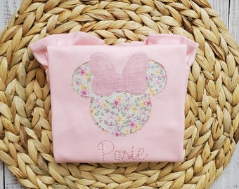 May include: A light pink short-sleeved shirt featuring a central applique of a mouse head silhouette with a bow. The silhouette is filled with a floral pattern of yellow, pink, and green flowers. The name Posie is embroidered in pink thread below.