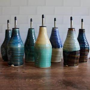 Op de afbeelding: Een verzameling van zeven keramische oliedispensers met gevarieerde glazuren in tinten blauw, groen, crème en bruin. Elke fles heeft een taps toelopende hals, een cilindrisch lichaam met subtiele horizontale ribbels en een zilverkleurige metalen schenktuit.