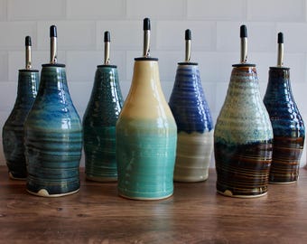 Puede incluir: Una colección de siete dispensadores de aceite de cerámica con acabados de esmalte variados en tonos azul, verde, crema y marrón. Cada botella presenta un cuello cónico, cuerpo cilíndrico con sutiles estrías horizontales y un vertedor de metal plateado.