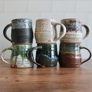 May include: Six handmade ceramic mugs in various colors and textures are stacked on a wooden surface. The mugs feature a variety of glazes, including speckled, dripped, and solid colors. Each mug has a large, curved handle.
