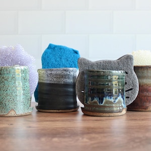 Op de afbeelding: Een verzameling handgemaakte keramische sponsenbakjes in verschillende kleuren en texturen. Elke houder is gecombineerd met een kleurrijke spons. De houders zijn in groen-, zwart- en bruintinten, met een katvormige houder.
