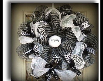 May include: A decorative wreath in black, white, and silver, featuring a basketball with the word "SPURS" and team logos. The wreath is made of mesh and ribbons, with a mix of solid and patterned elements.