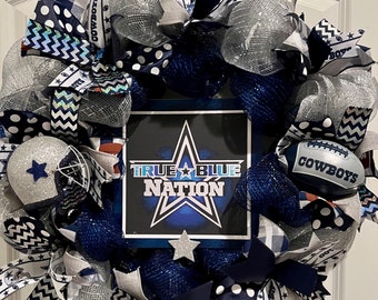May include: A decorative wreath with a football theme. The wreath is decorated with navy blue, silver, and white ribbons, a football, and a helmet. The centrepiece features a sign with the text "TRUE BLUE NATION" and a star.
