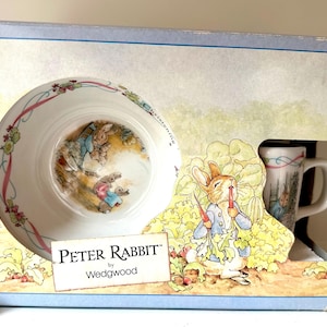 May include: A Peter Rabbit by Wedgwood boxed set featuring a white ceramic bowl and mug with illustrations of Peter Rabbit and other characters from the book. The box is decorated with a colourful illustration of Peter Rabbit in a garden.