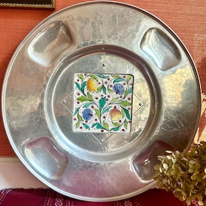 Könnte beinhalten: Ein großer, runder, gehammerter Metallteller mit einem quadratischen Mittelkachel mit einem Blumendesign mit gelben und pfirsichfarbenen Früchten und blauen Blumen. Der Teller hat vier vertiefte Bereiche.
