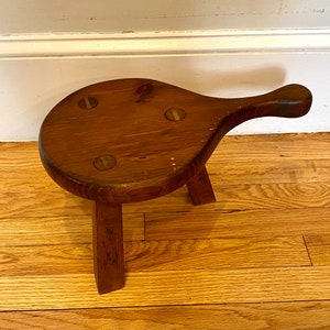 May include: A wooden stool with a rounded top and a single handle. The stool has three legs and is made of dark brown wood. There are three metal accents on the top of the stool.