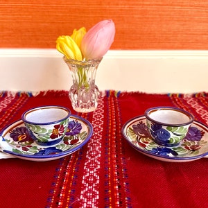May include: Two ceramic candle holders with blue and white floral designs. Each holder has a small cup for a candle and a handle. The holders are on a red and white striped table runner.
