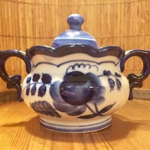 Pode incluir: Um açucareiro de porcelana branca com um design floral azul e branco. O açucareiro tem uma tampa e duas alças.