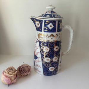 May include: A vintage porcelain coffee pot with a blue and gold floral design. The pot has a white background with a blue and gold floral pattern. The handle is white and the lid is white with a blue and gold floral pattern.