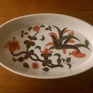 Puede incluir: Plato para hornear ovalado de cerámica blanca con un diseño floral en tonos verdes, naranjas y marrones. El diseño presenta hojas, flores y tallos estilizados.