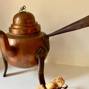 May include: A copper teapot with a rounded body, a curved spout, and a domed lid featuring a small metal loop handle. The teapot is supported by three dark metal legs and includes a long, straight, dark metal handle extending from the side.