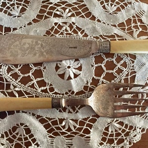 Puede incluir: Un juego de cubiertos de plata antiguos con mangos de marfil. El cuchillo tiene un diseño ornamentado y el tenedor tiene un diseño simple. Los cubiertos están sobre un encaje blanco.