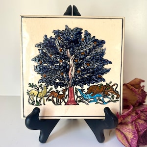 May include: A ceramic tile with a hand-painted design of a tree with blue leaves and orange fruit.  Animals surround the base of the tree, including a deer, a lion, and a bird.