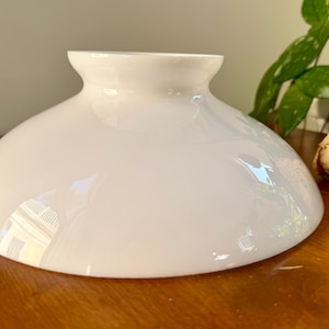 May include: A white, glossy glass lampshade with a rounded, bowl-like shape. The lampshade has a flared base and a narrower, slightly curved top. The surface reflects light, and it sits on a wooden surface.