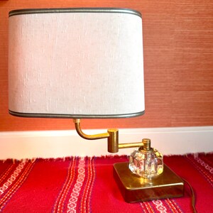 May include: A vintage table lamp with a rectangular off-white lampshade, trimmed with a dark green border. The lamp features a brass arm and base, and a clear, faceted crystal knob. The lamp sits on a red patterned rug.