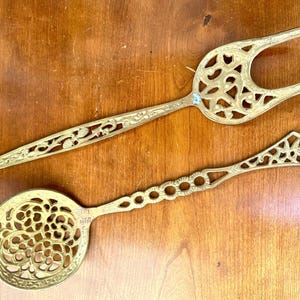 Op de afbeelding: Twee antieke messing serveerbestekken: een vork en een lepel. De vork heeft een lange steel met bloemendetails en een decoratief opengewerkt ontwerp. De lepel heeft een ronde kom met een opengewerkt bloemmotief.