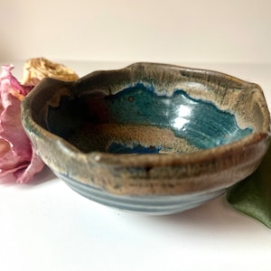 May include: A small, handmade ceramic bowl with a unique, irregular rim. The bowl features a blend of colors, including shades of blue, brown, and tan, creating a natural, earthy aesthetic. The bowl is likely suitable for serving small portions of food.
