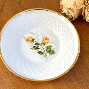 May include: White porcelain saucer with a gold rim and a raised diamond pattern. The center features a smaller, matching dish with a floral design of yellow roses and green leaves. A decorative piece for serving.