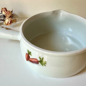Puede incluir: Cuenco de sopa de cerámica blanca con asa y boquilla. El cuenco presenta un diseño pintado de zanahorias y verduras. El cuenco está vacío y sobre una superficie blanca.