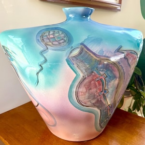 May include: A large, decorative ceramic vase with a wide, angular body and a narrow neck. The vase features a colorful abstract design in shades of blue, pink, and purple, with a globe-like motif. The vase is displayed on a wooden table.