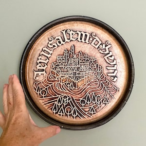 Può includere: Un piatto rotondo in ceramica con una glassa marrone e un disegno bianco di uno skyline di città con le parole "Gerusalemme" in ebraico e inglese.