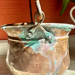May include: A vintage copper bucket with a metal handle and a patina finish. The bucket has a rounded bottom and a wide opening. The bucket is made of copper and has a green patina finish.